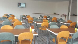 Ein Klassenzimmer mit Reihen von Tischen und Stühlen, einem Fernseher an der Wand und einem Regal mit Gegenständen.