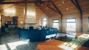 A spacious room with a high ceiling, wooden beams, a stone fireplace, a blue sofa, a dining table with placemats, and a view through large windows. - Mandala Bliss Mongolia (Ulaanbaatar)
