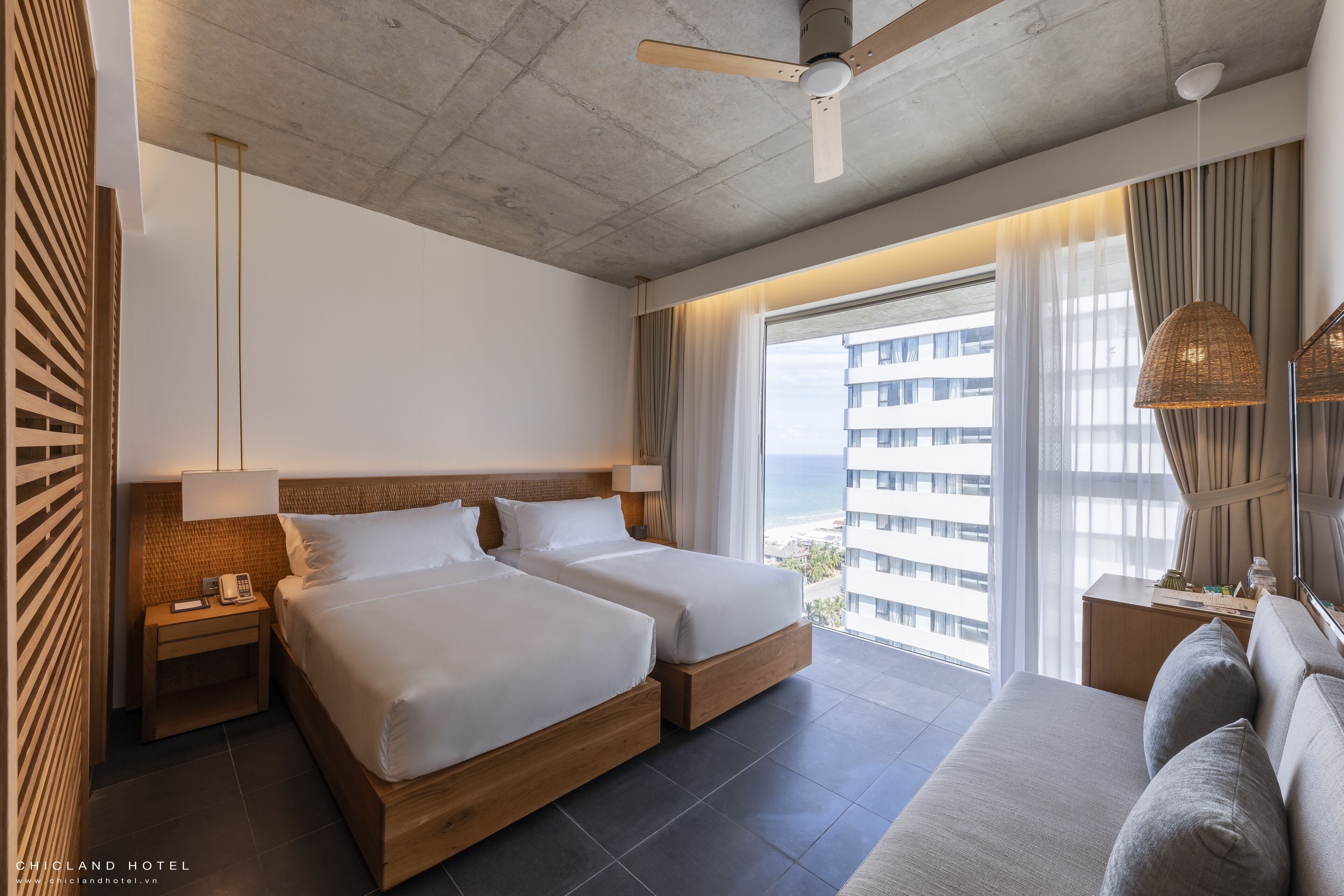 Ein Hotelzimmer mit zwei Betten, einem Deckenventilator, einem großen Fenster mit Vorhängen und Blick auf die Stadt.