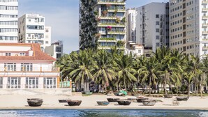 Um edifício alto com varandas, uma praia com palmeiras e um corpo d'água em primeiro plano.