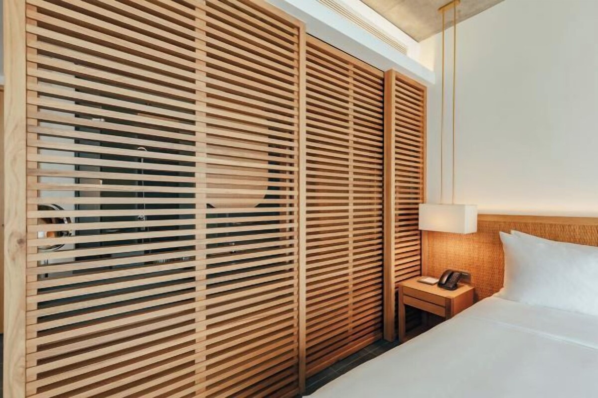 A hotel room with a bed, bedside table, lamp, and wooden blinds.