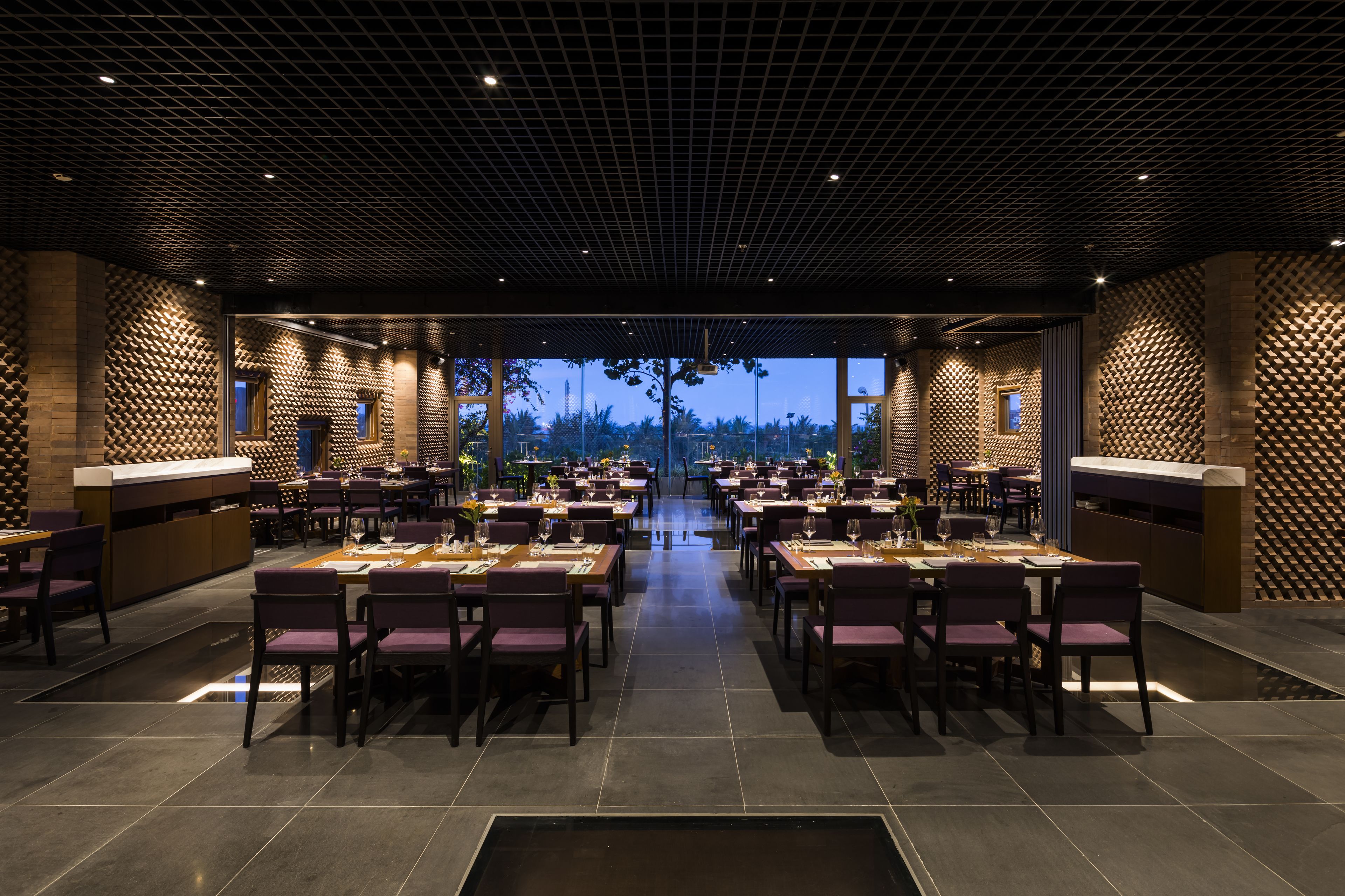 Un restaurante espacioso de diseño moderno, con una zona de comedor central con numerosas mesas y sillas, y grandes ventanales que ofrecen vistas al exterior.