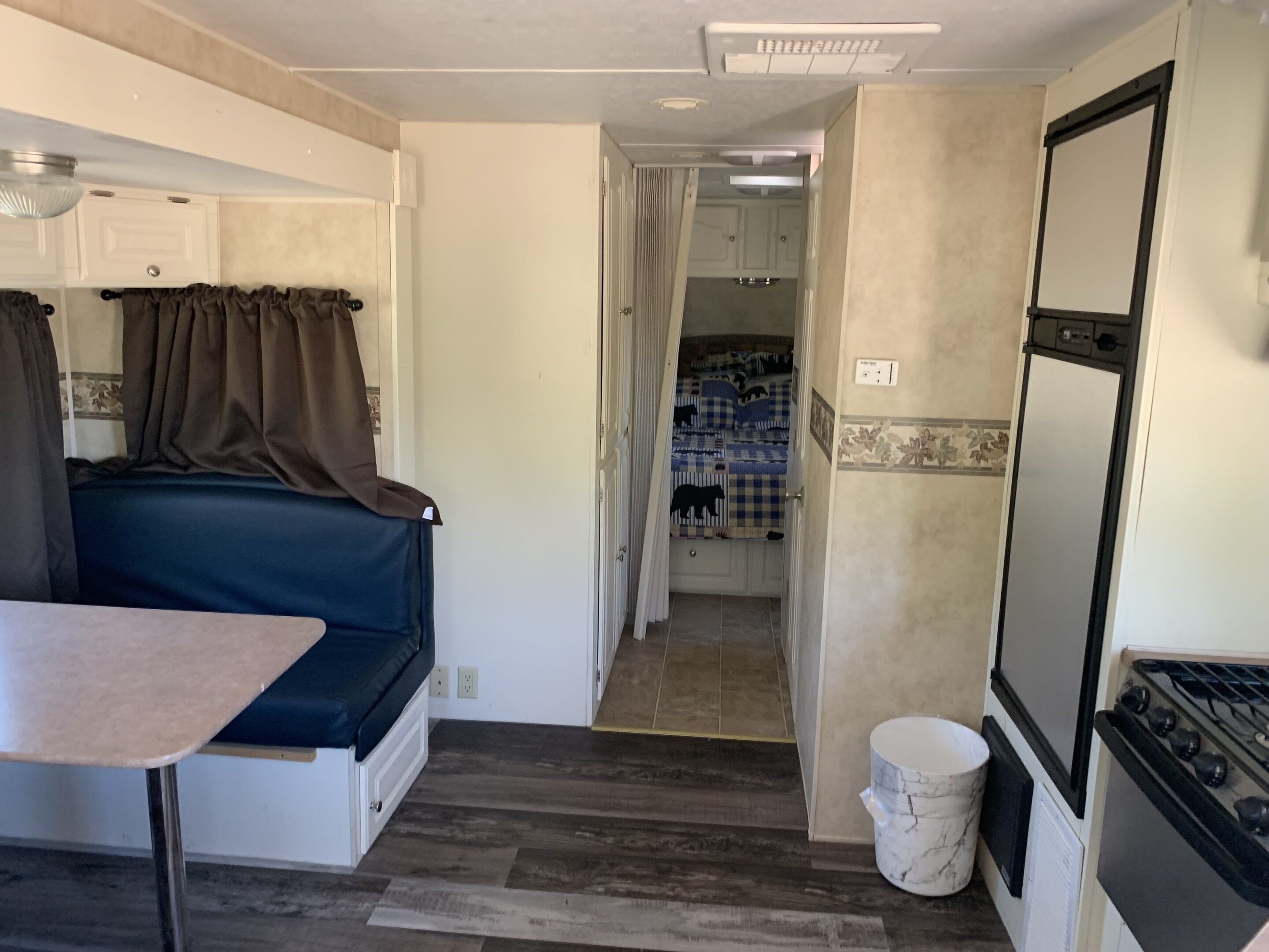 A compact RV interior with a kitchenette, a small dining area, and a bedroom with a bed and curtains.