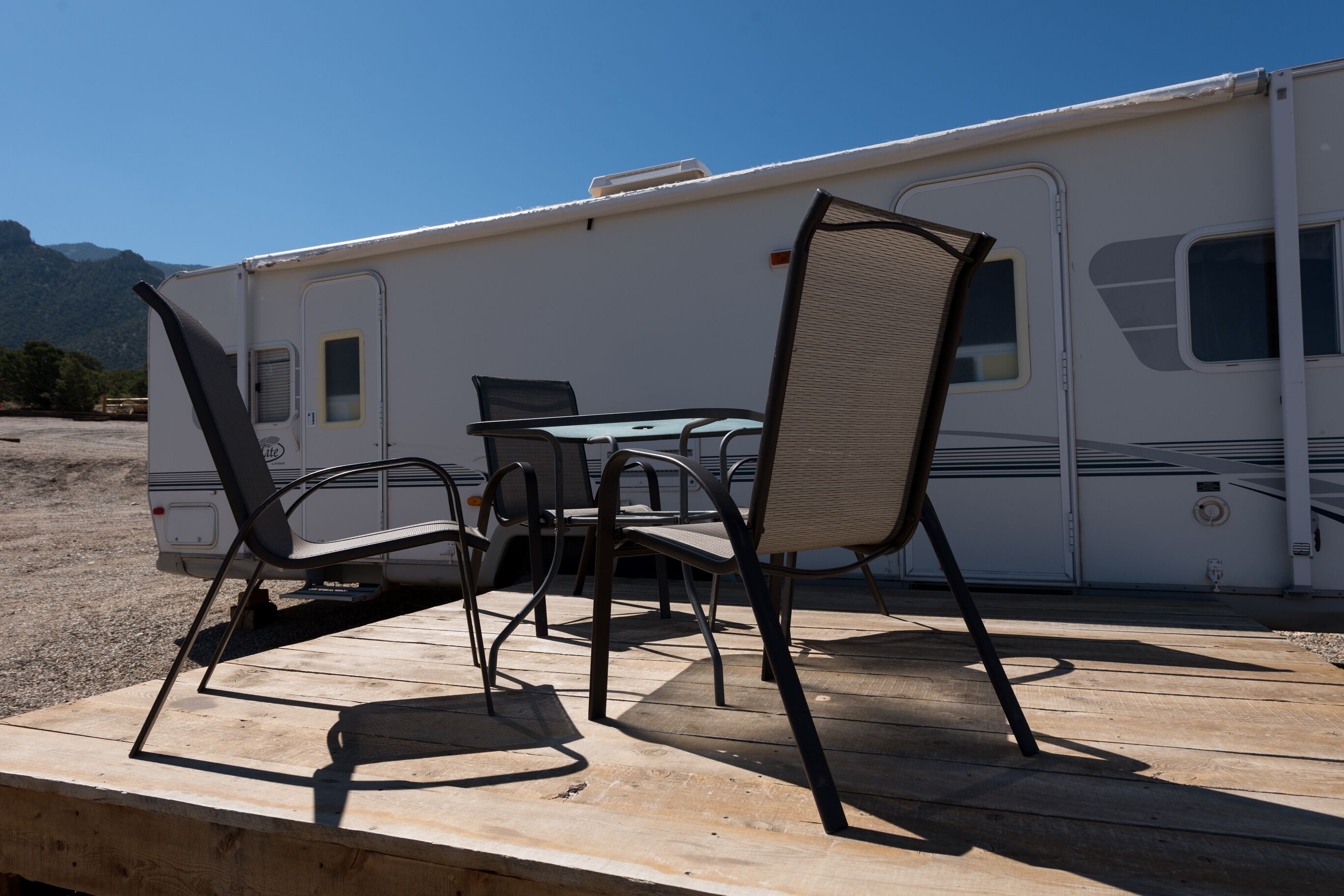Ein Campingplatz mit Esstisch und Stühlen auf einer Holzterrasse, einem weißen Wohnmobil und einer Bergkulisse.