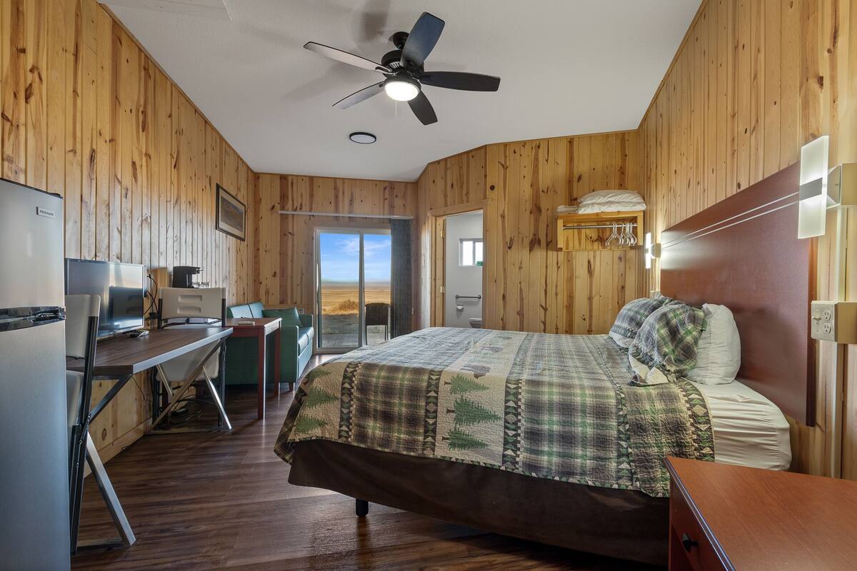 A bedroom with a bed, a desk, a chair, a ceiling fan, and a view of the outdoors.