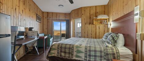 A bedroom with a bed, a desk, a chair, a ceiling fan, and a view of the outdoors.