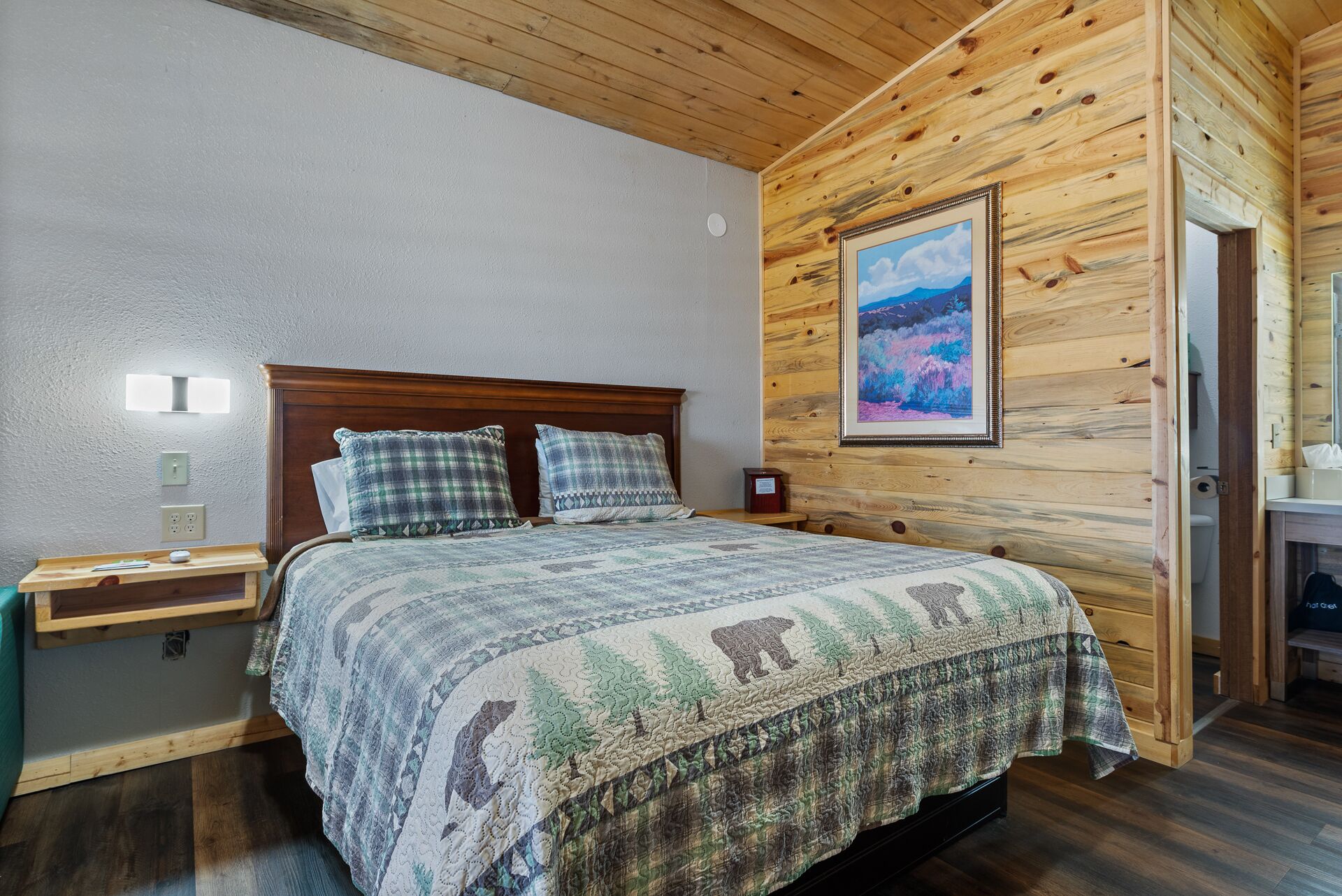 Une chambre avec un plafond en bois, un lit recouvert d’une couvre-lit à motifs, deux oreillers, une table de chevet, une lampe fixée au mur et un tableau encadré accroché au mur.