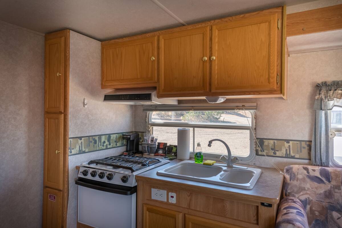 A compact RV kitchen with wooden cabinets, a stove, a sink, and a window providing a view of the outdoors.