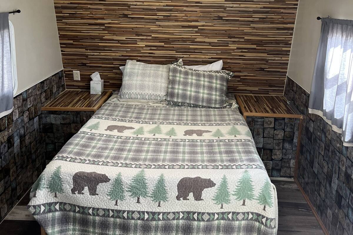 A bedroom with a wooden headboard, a bed with a forest-themed bedspread, two bedside tables, and a window with curtains.