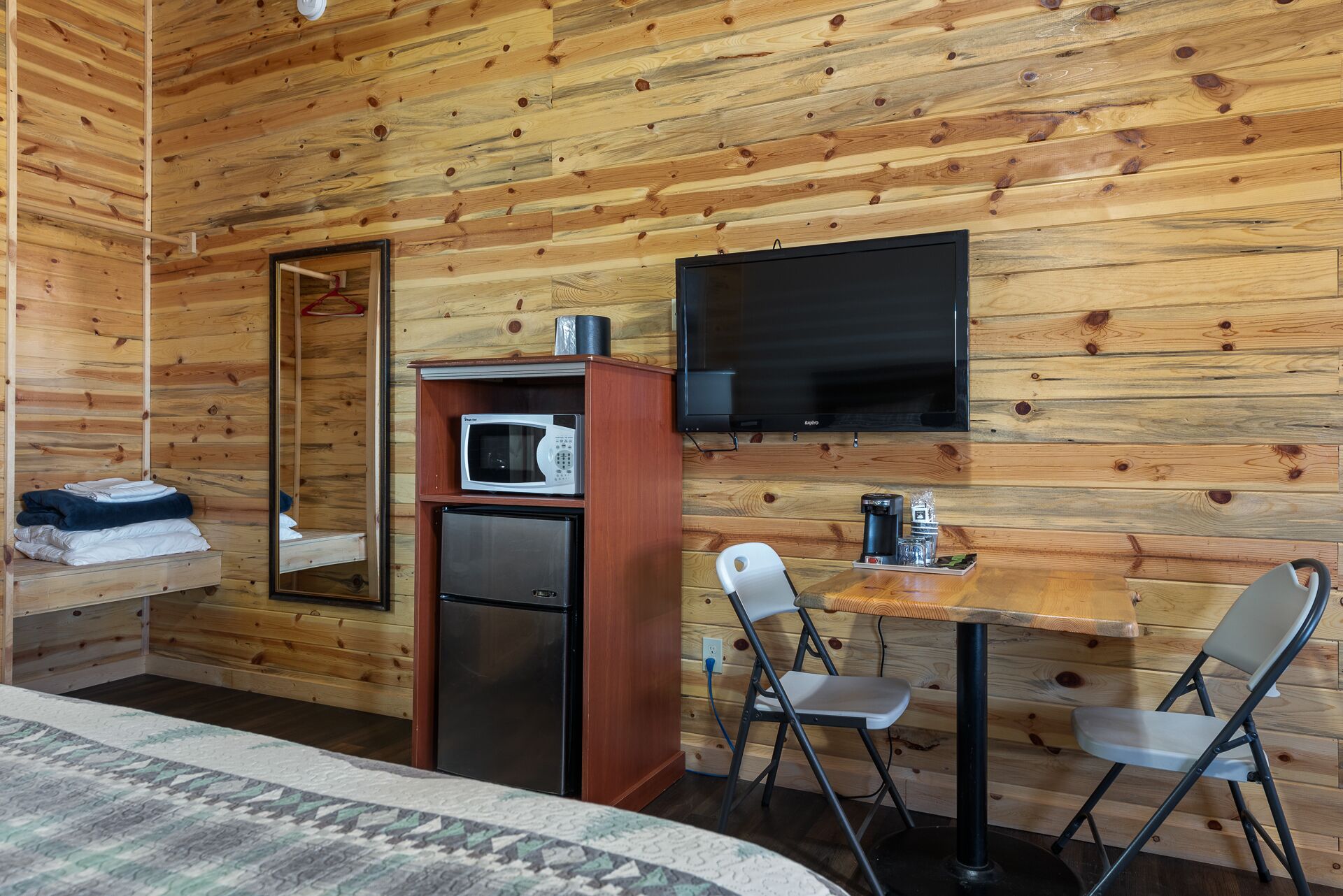 A compact room with a bed, a small kitchenette with a microwave and refrigerator, a TV mounted on the wall, and two foldable chairs at a small table.