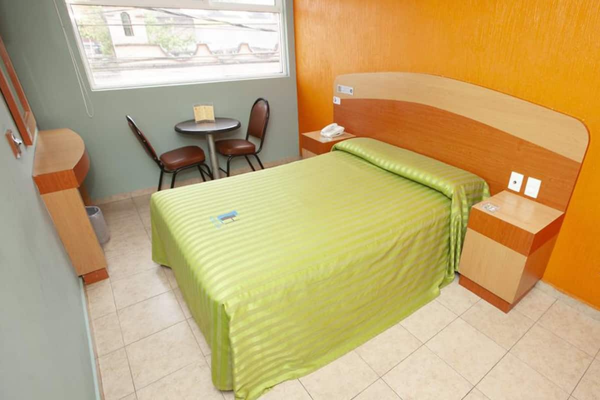 Une chambre d’hôtel avec un lit, une petite table, deux chaises, une fenêtre et une tête de lit en bois.