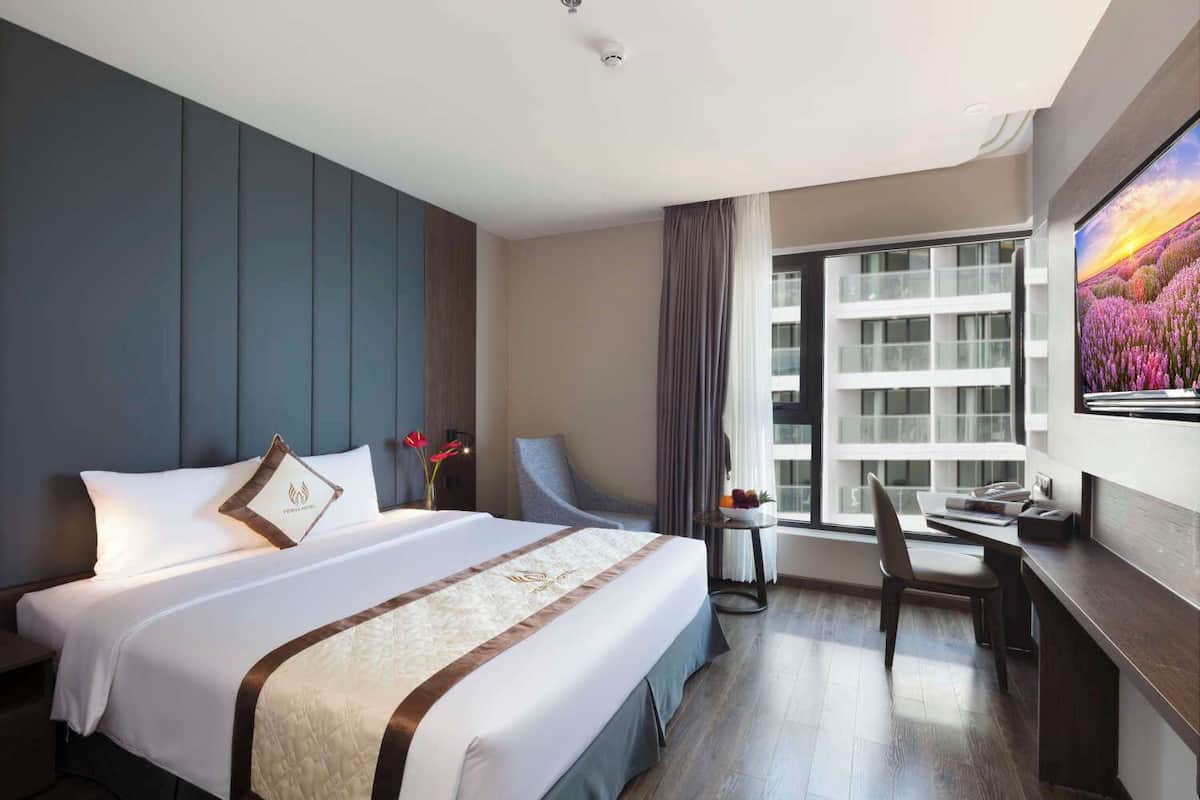 A modern hotel room with a large bed, a desk, and a view of a cityscape through the window.