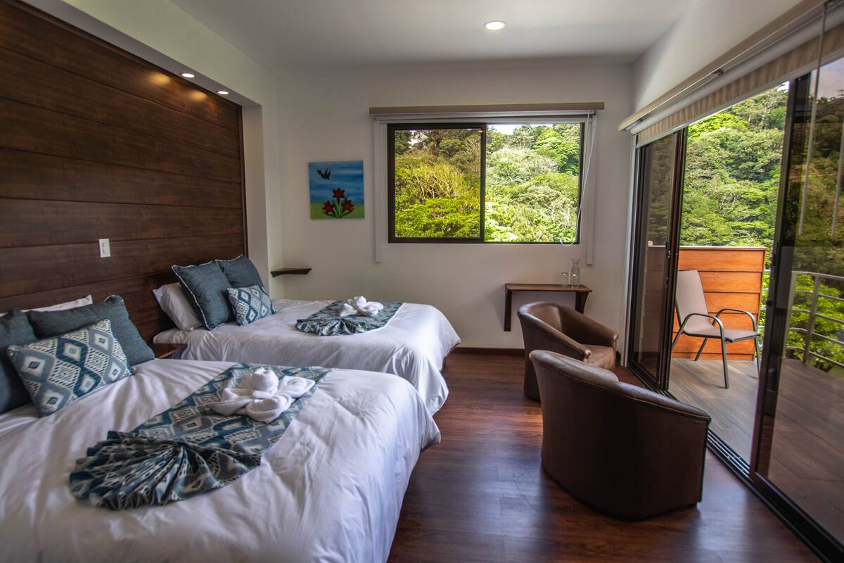 A hotel room with two beds, a small table, a chair, and a large window offering a view of greenery.