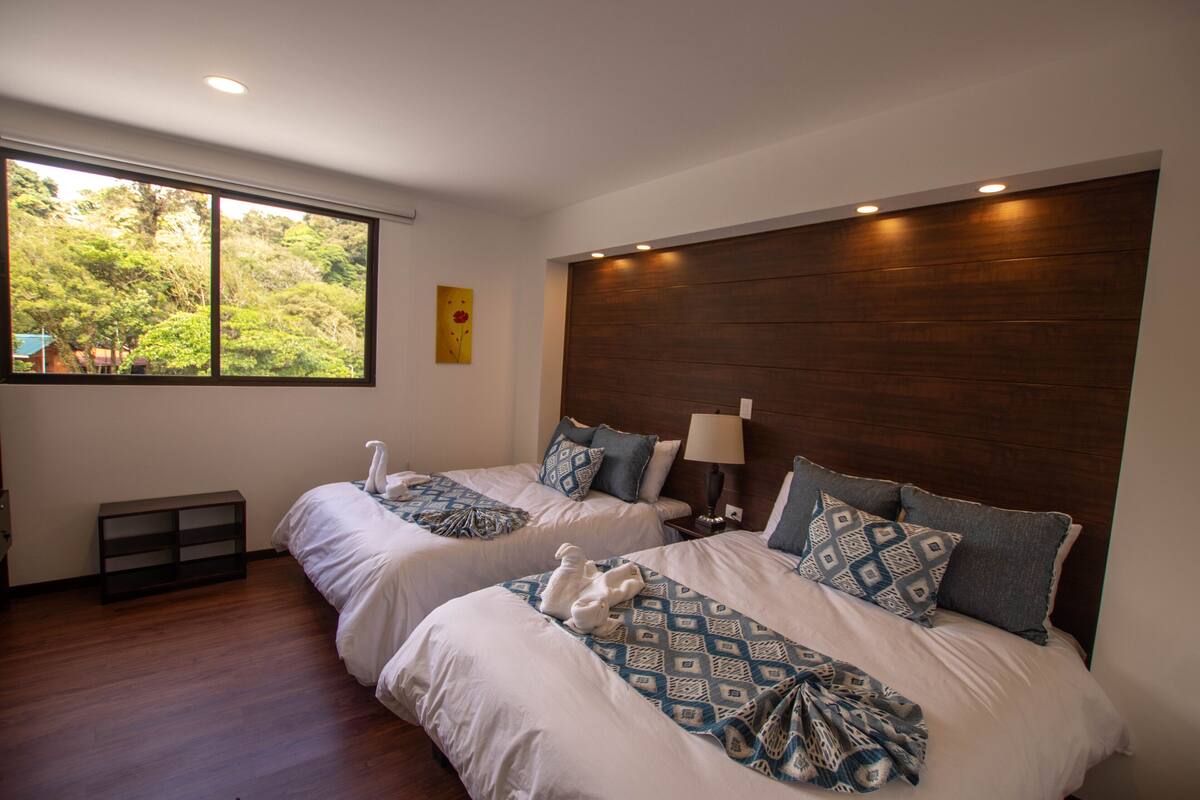 A hotel room with two beds, a wooden headboard, a window with a view of trees, and a small shelf.