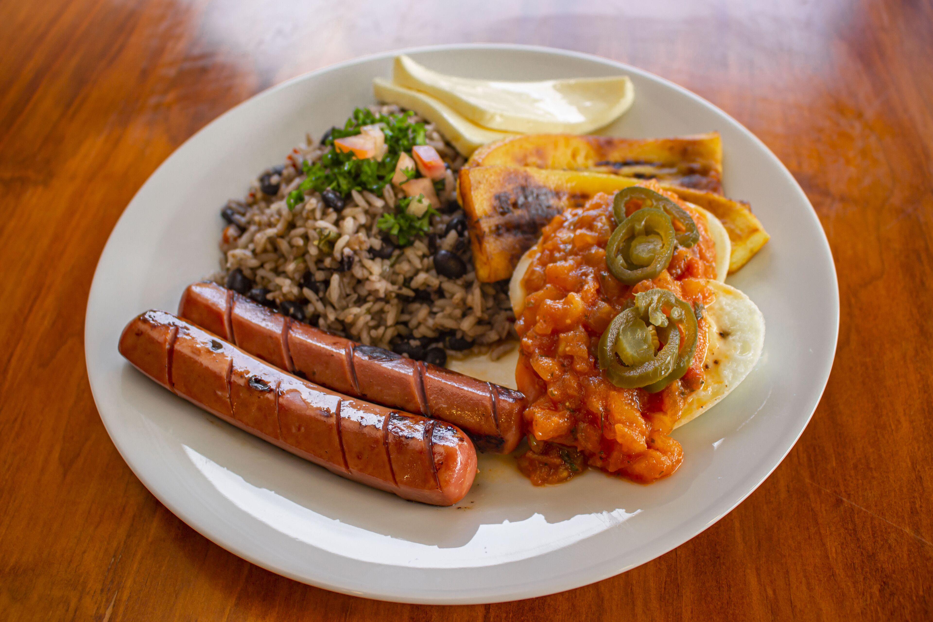 A plate of food including rice, black beans, two sausages, a slice of cheese, a slice of bread, and a slice of plantain.