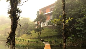 Une jetée en bois mène à un bâtiment à l’architecture moderne, entouré d’une végétation luxuriante et bordé par une étendue d’eau calme.