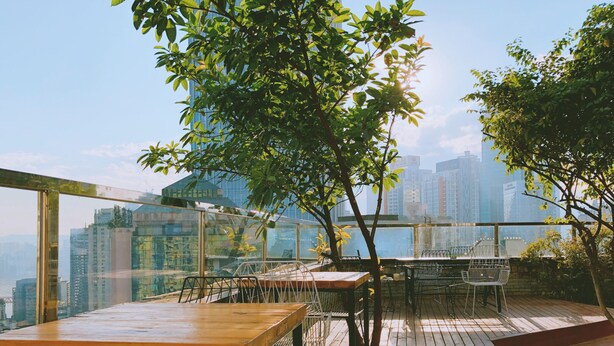 An outdoor dining area with wooden tables and chairs, surrounded by glass railings, overlooking a cityscape.