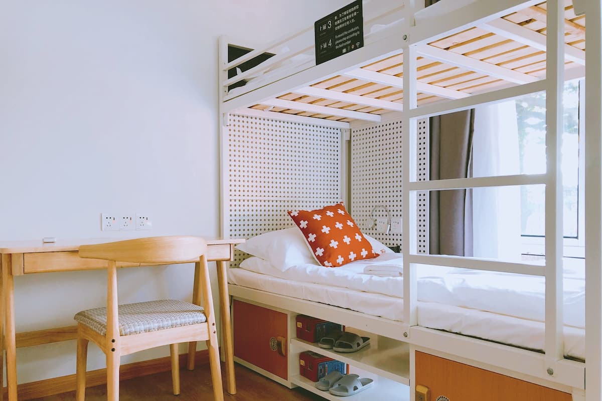 A bunk bed with a desk and chair, a window with curtains, and a bed with a red pillow.