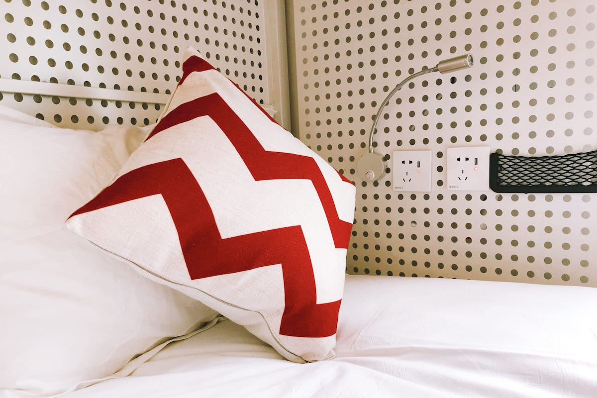 A bed with a white sheet and a red and white pillow. There is a power outlet and a plug on the wall.