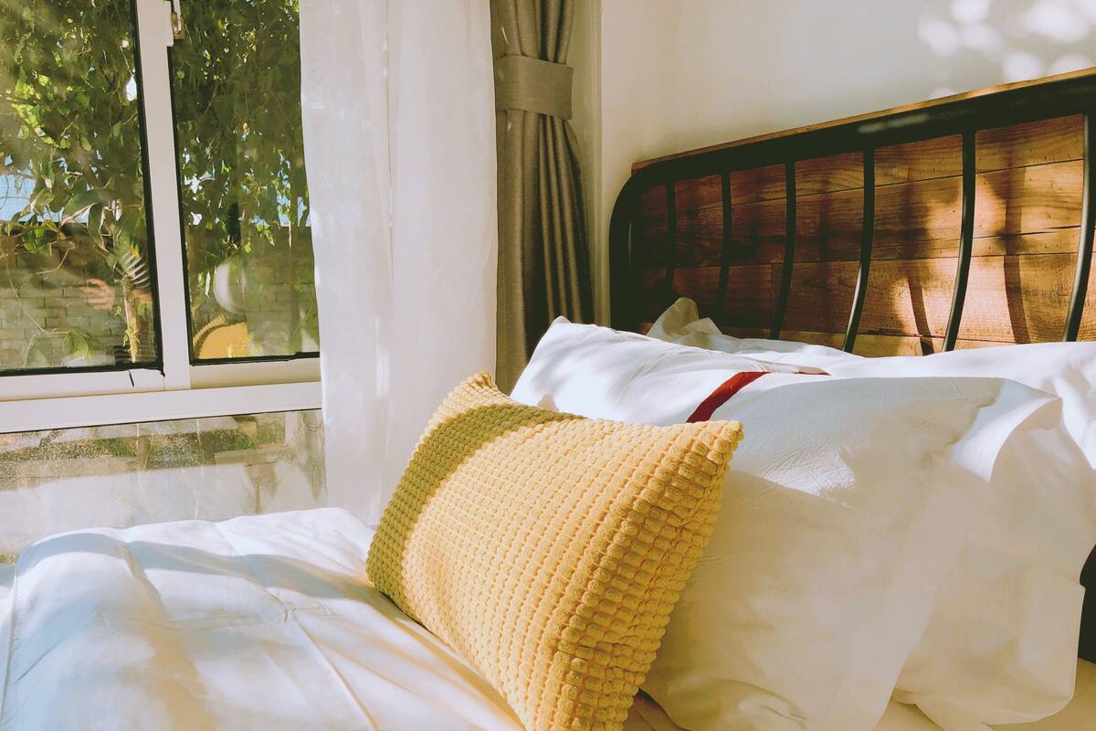 A bed with white linens and a yellow textured pillow, a wooden headboard, and a window with curtains.