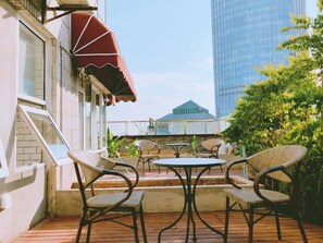 Une terrasse extérieure avec des chaises et une table en osier, un auvent rouge et une vue sur un immeuble de grande hauteur.