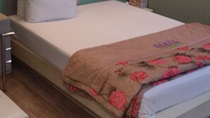 Una cama individual con colchón blanco y manta estampada, una almohada y una mesita de noche con lámpara. La habitación tiene paredes verdes y suelo de madera.