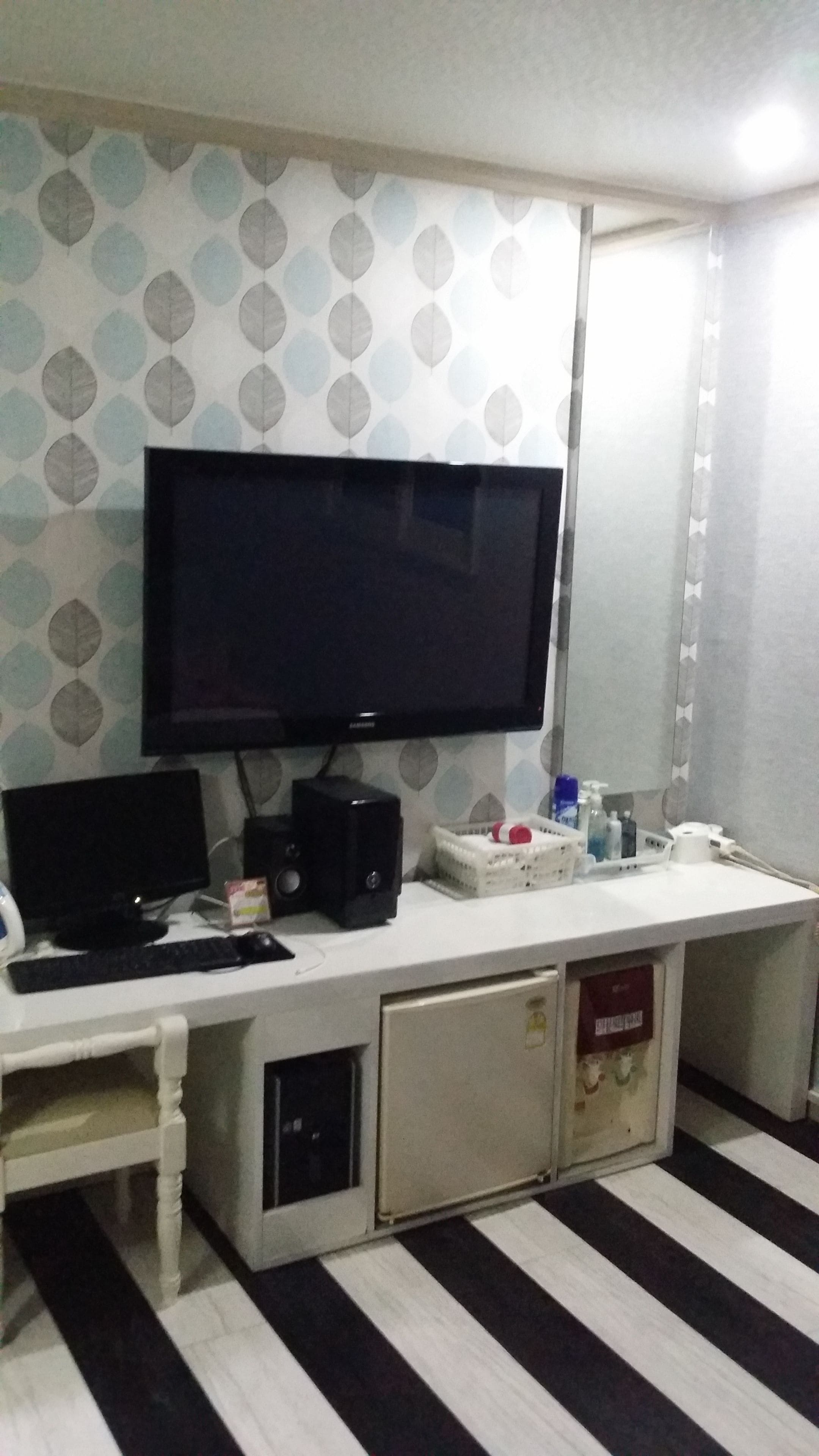 A room with a large flat-screen TV mounted on the wall, a desk with a computer, keyboard, and mouse, and a small refrigerator.