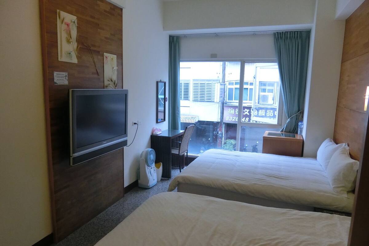 A hotel room with two beds, a TV, a fan, and a view of the city through the window.
