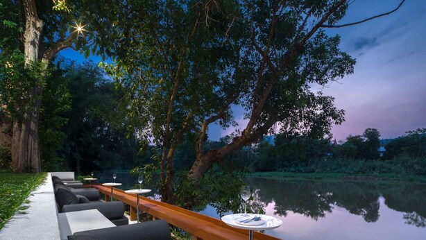 A serene outdoor seating area with modern sofas and tables by a reflective water body, surrounded by lush greenery and trees. - Little Shelter Hotel Chiangmai  (Chiang Mai)