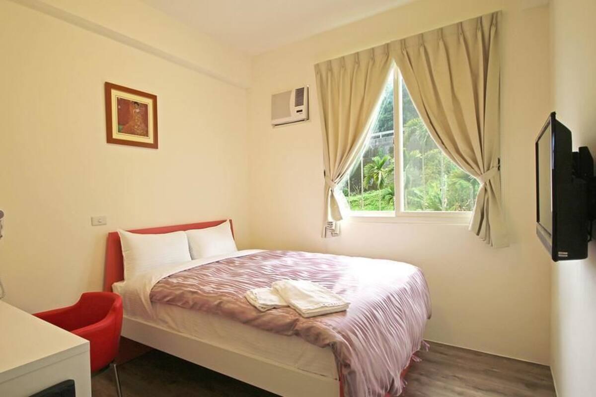 A bedroom with a bed, a desk, a chair, a window with curtains, a picture on the wall, and an air conditioning unit.