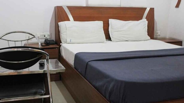 A hotel room with a wooden headboard, white pillows, a dark blue bedspread, a small bedside table with a lamp, and a chair. - FabHotel My Choice Abids (Hyderabad)
