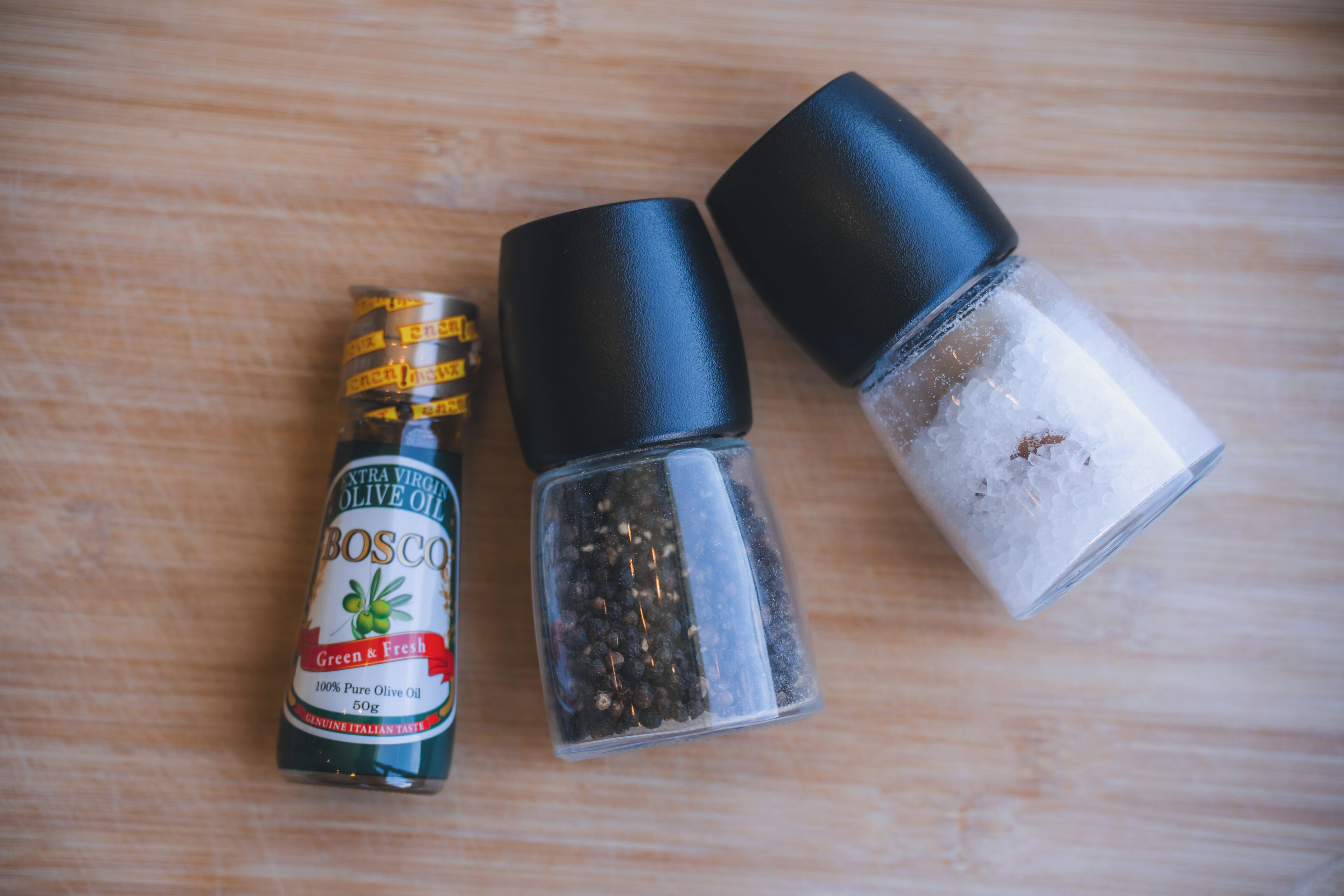 A bottle of extra virgin olive oil, a pepper grinder, and a salt shaker on a wooden surface.