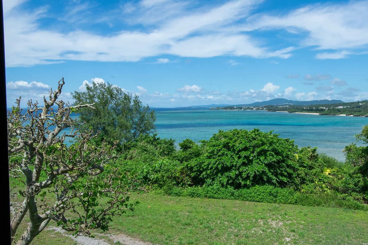A coastal landscape with a clear view of the sea, lush greenery, and distant hills.