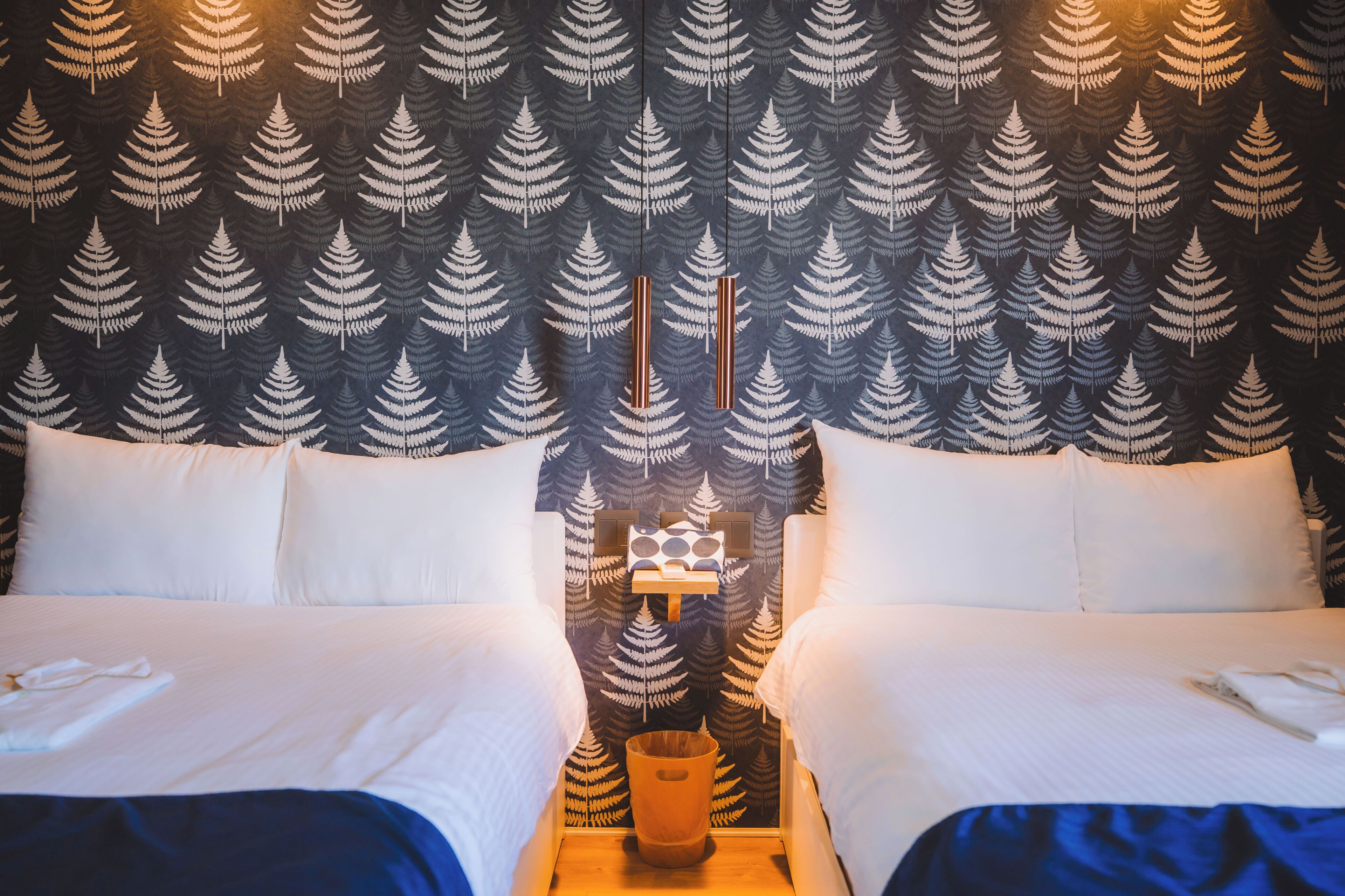 A hotel room with a bed, two pillows, a bedside table with a lamp, and a wall with a fern pattern.