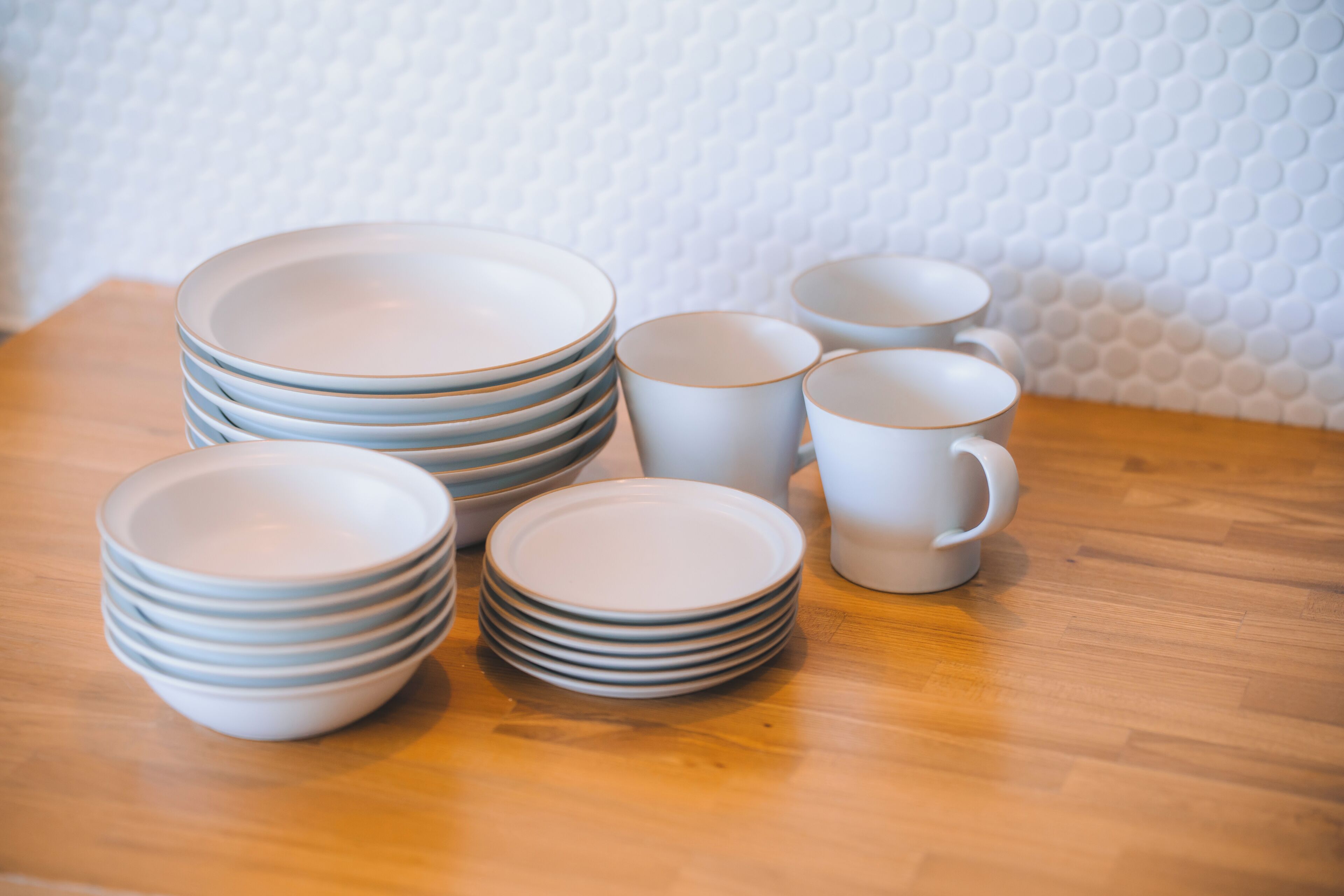 A stack of white plates and bowls on a wooden table with a textured white wall in the background.