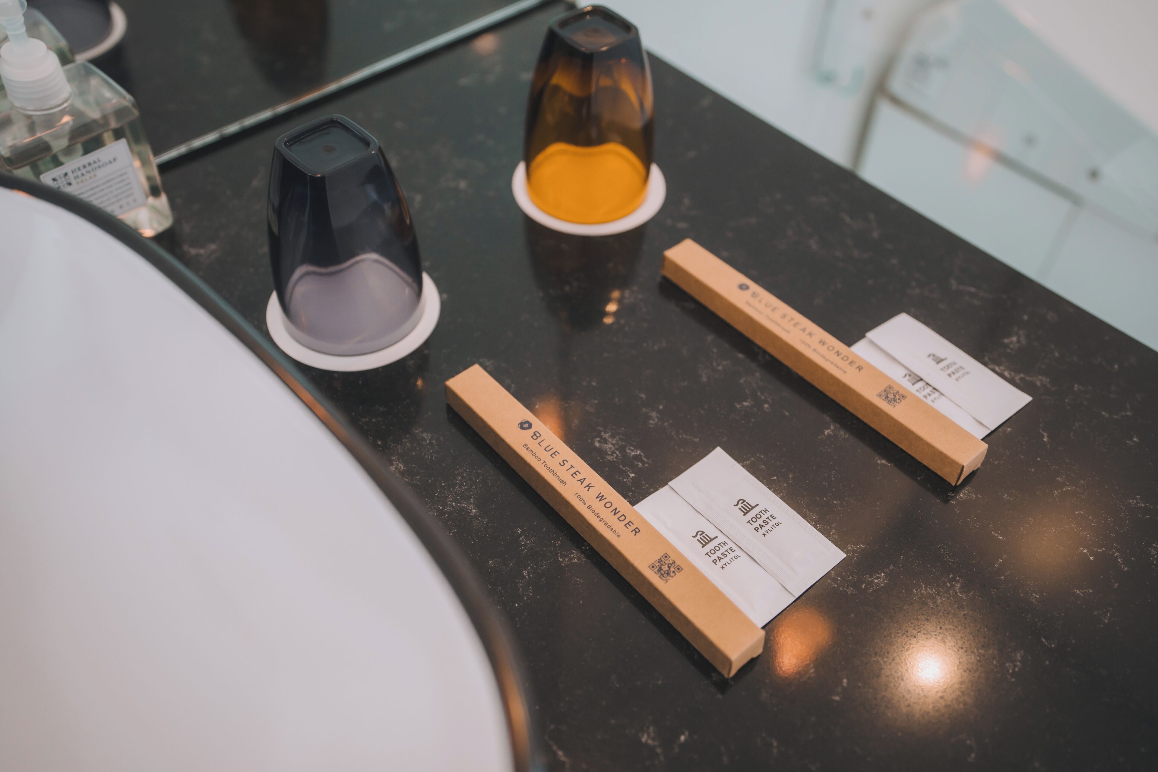 A bathroom counter with a mirror, a bottle of hand soap, two toothbrushes, and two boxes of toothpaste.