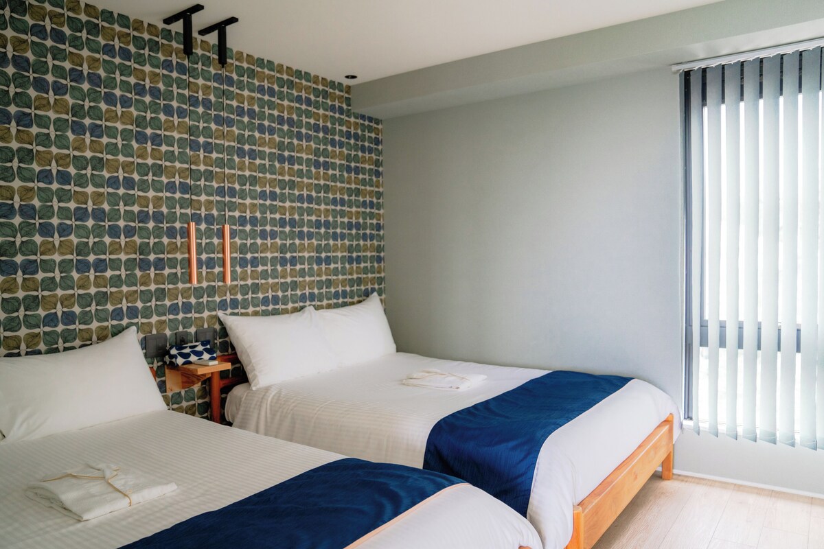 A hotel room with two beds, a patterned wall, a nightstand, and a window with blinds.