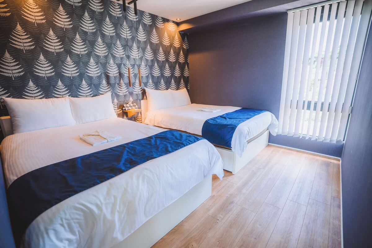 A hotel room with two beds, a patterned wall, a window with blinds, and a wooden floor.