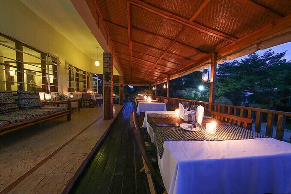 A covered outdoor dining area with tables set for dinner, a sofa, and a view of the surrounding trees.