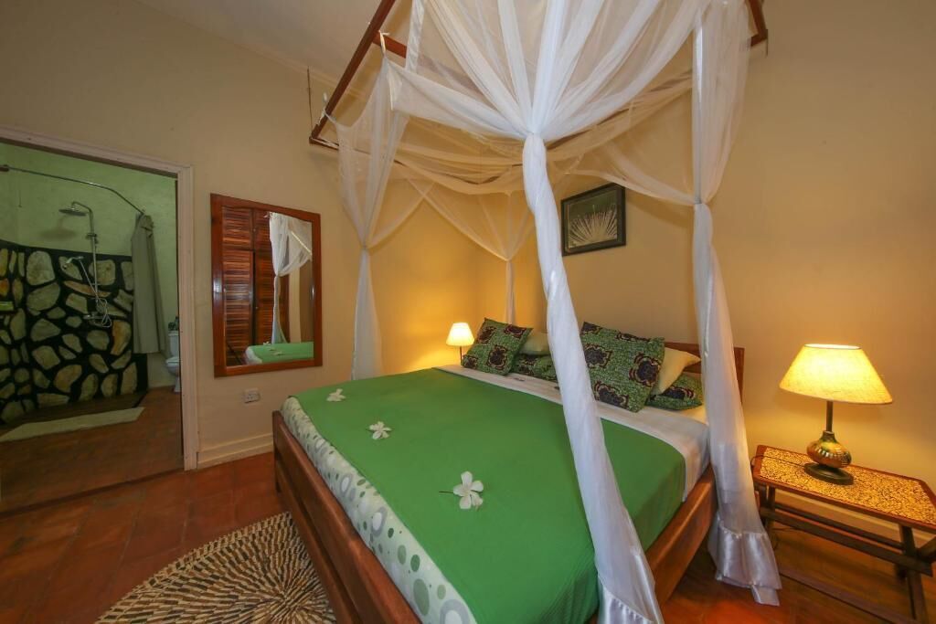 A room with a four-poster bed, a nightstand with a lamp, a mirror, and a green bedspread.