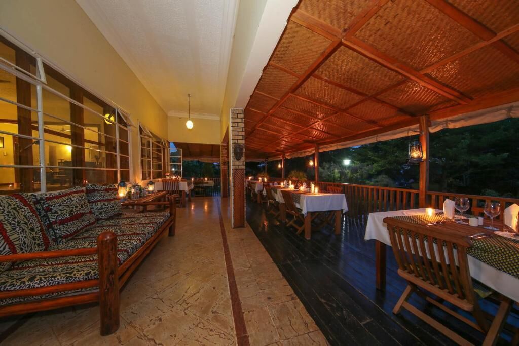 An outdoor dining area with wooden furniture and tables, a covered section with a woven roof, and a view of the outdoors.