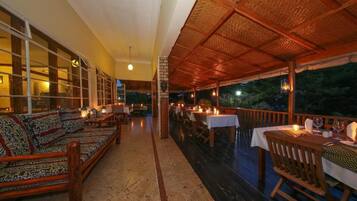 An outdoor dining area with wooden furniture and tables, a covered section with a woven roof, and a view of the outdoors.