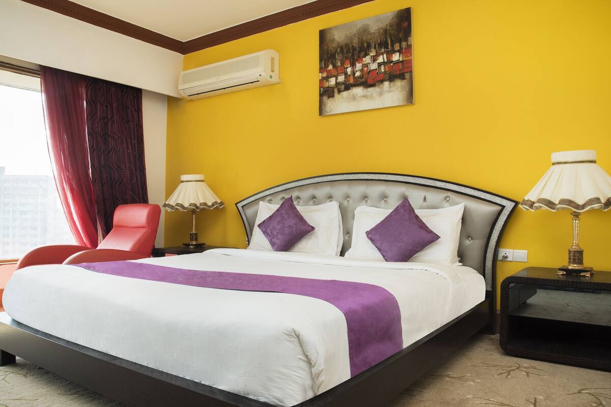 A hotel room with a bed, a red chair, a nightstand, an air conditioner, and a painting on the wall.
