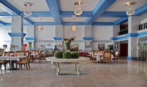 A spacious hotel lobby with high ceilings, blue accents, and chandeliers. Tables and chairs are arranged for dining or meetings. - Treebo Tryst Royal Palm (Mumbai)