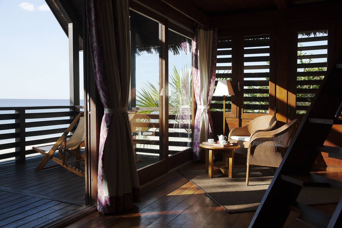 A wooden deck with a rocking chair, a table with a lamp, and a view of the ocean through large windows.