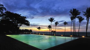 Een overloopzwembad met een houten terras, palmbomen en uitzicht op een zonsondergang.