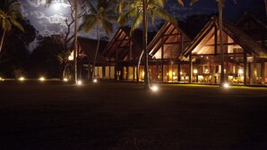 Een resort met rieten gebouwen, palmbomen en een nachthemel met een zichtbare maan.