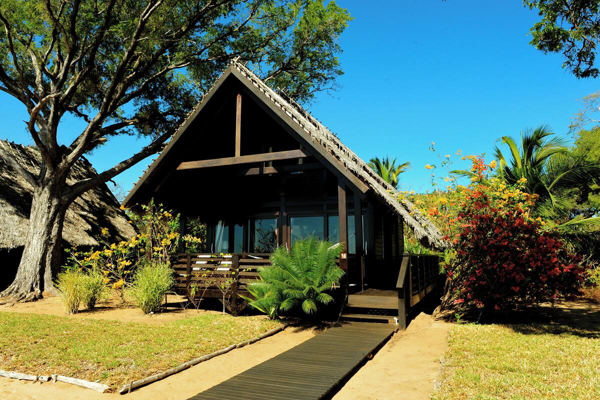 A wooden lodge with a thatched roof, surrounded by tropical vegetation and a clear blue sky.