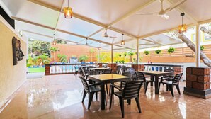 Un área de comedor al aire libre techada con mesas y sillas, ventiladores de techo y plantas colgantes.