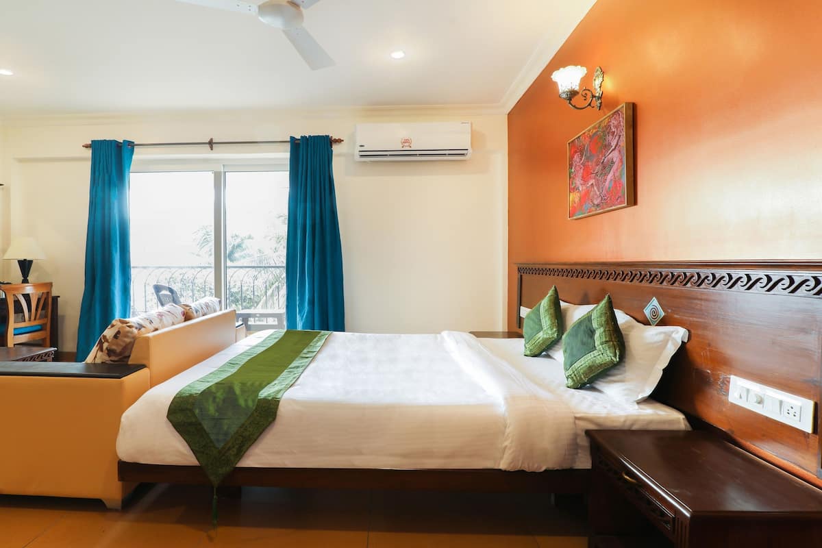 A hotel room with a bed, a desk with a chair, a window with curtains, an air conditioning unit, and a painting on the wall.